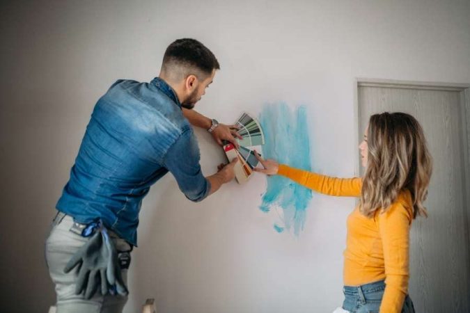 Couple comparant des échantillons de couleurs sur un mur avec une touche de peinture bleue, pour choisir la bonne peinture de leur intérieur.